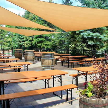 L'area esterna di Elm Creek Brewing presenta diversi set da birreria Classic in colore pino con telai neri, disposti sotto grandi vele ombreggianti. L'area è incorniciata da alberi verdi.